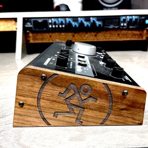 May include: Side view of a black audio interface with a wooden side panel. The panel has a logo of a stylized person running with the letters "M" and "A" on either side. The interface has a silver knob and several black knobs.