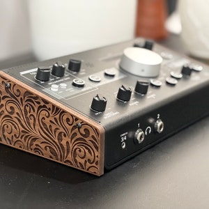 Op de afbeelding: Zwart audio-interface met meerdere knoppen, draaiknoppen en ingangsconnectoren. Het apparaat heeft een zilveren bedieningsknop en een houten paneel met een gegraveerd bloemmotief. De tekst "STEREO 3/4" is zichtbaar.