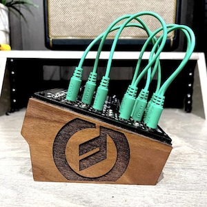 以下が含まれることがあります： レーザー彫刻ロゴが入った木製の電子音楽モジュール。前面パネルには複数の緑色のパッチケーブルが接続されています。モジュールは角度がついており、そのデザインと接続されたケーブルを示しています。