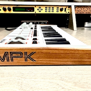 May include: A white and black Akai MPK MIDI keyboard with a wooden base. The keyboard has black and white keys, white knobs, and red buttons. The letters "MPK" are etched into the wooden base.