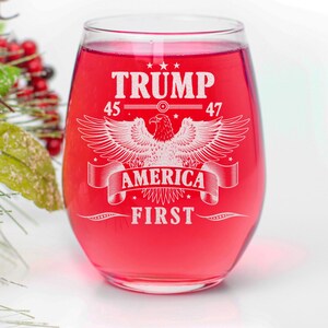 Puede incluir: Una copa de vino tinto con un dise&ntilde;o blanco que dice "TRUMP 45-47 AMERICA FIRST" con un &aacute;guila en el centro.