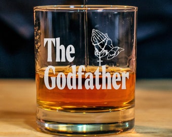 May include: A clear glass with the text "The Godfather" and a praying hands design is being filled with amber liquid from a bottle.