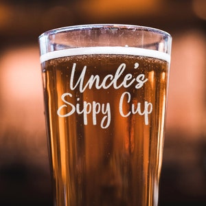 May include: A tall glass beer mug with a clear glass base and a frosted glass top. The mug is filled with a golden amber beer and has the text "Uncle's Sippy Cup" etched into the side.