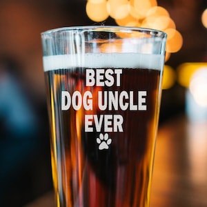 May include: A clear pint glass filled with a dark amber liquid, likely beer, with a foamy head. The glass has the words "BEST DOG UNCLE EVER" in white, along with a paw print. The glass sits on a wooden surface.