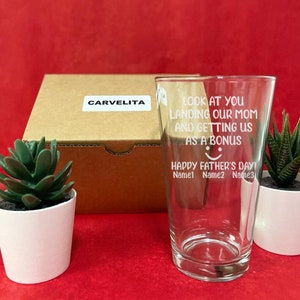 May include: A clear glass with a white inscription that reads "Look at you landing our mom and getting us as a bonus Happy Father's Day! Name1 Name2 Name3". The glass is sitting on a table with a brown box and two potted succulents.