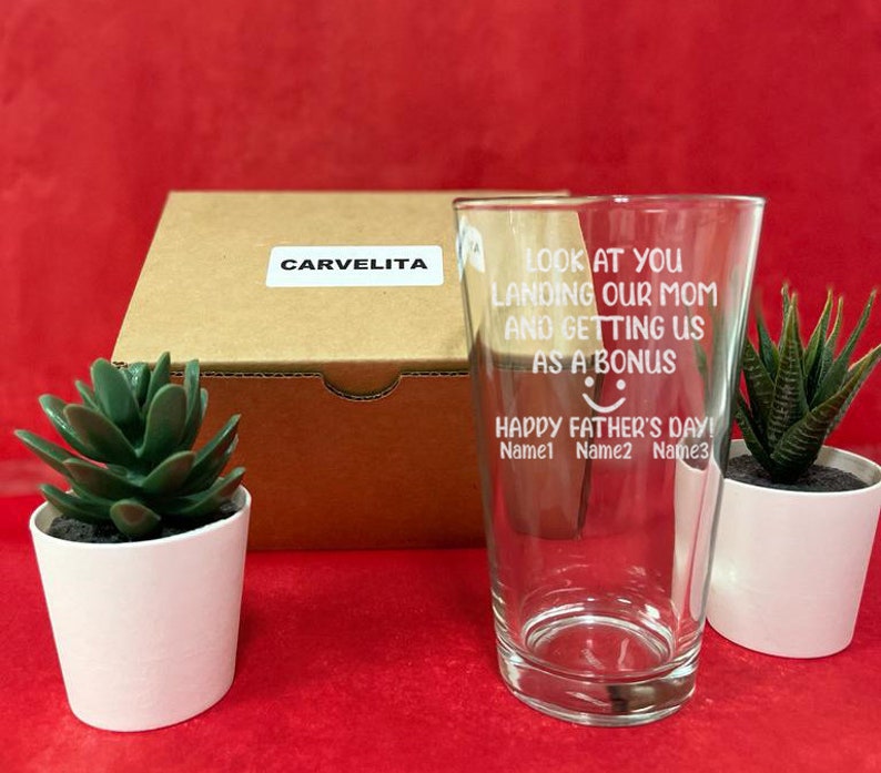 May include: A clear glass with a white inscription that reads "Look at you landing our mom and getting us as a bonus Happy Father's Day! Name1 Name2 Name3". The glass is sitting on a table with a brown box and two potted succulents.