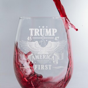 Puede incluir: Una copa de vino de vidrio transparente con un dise&ntilde;o grabado que presenta un &aacute;guila, el texto "TRUMP 45 47 AMERICA FIRST" y estrellas. Se vierte vino tinto en la copa.
