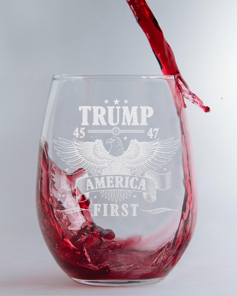 Puede incluir: Una copa de vino de vidrio transparente con un dise&ntilde;o grabado que presenta un &aacute;guila, el texto "TRUMP 45 47 AMERICA FIRST" y estrellas. Se vierte vino tinto en la copa.
