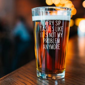 Pode incluir: Um copo de cerveja cheio de cerveja, com o texto branco "EVERY SIP TASTES LIKE IT'S NOT MY PROBLEM ANYMORE". O copo est&aacute; sobre uma superf&iacute;cie de madeira, com um fundo desfocado.