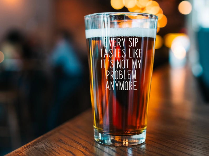 Pode incluir: Um copo de cerveja cheio de cerveja, com o texto branco "EVERY SIP TASTES LIKE IT'S NOT MY PROBLEM ANYMORE". O copo est&aacute; sobre uma superf&iacute;cie de madeira, com um fundo desfocado.
