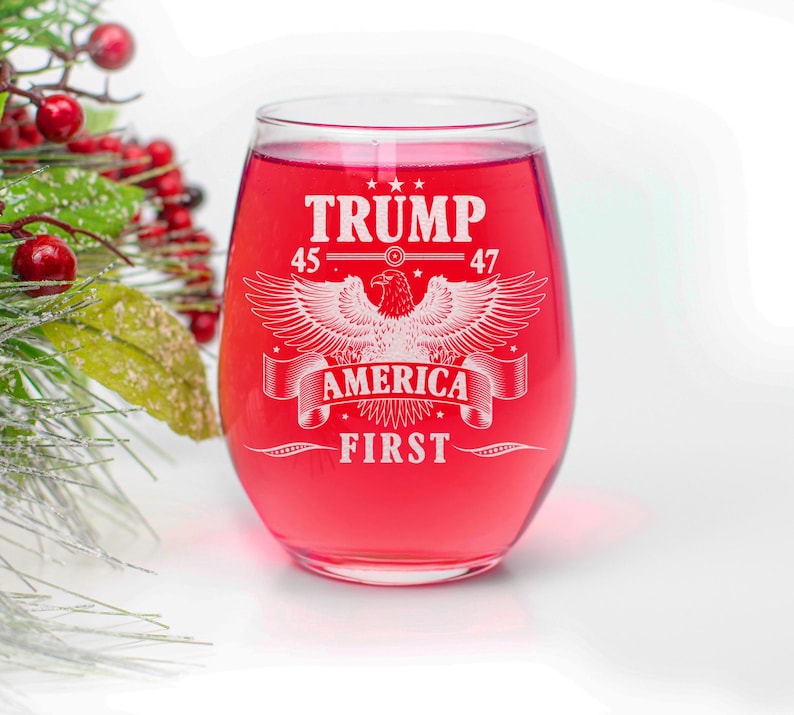 Puede incluir: Una copa de vino tinto con un dise&ntilde;o blanco que dice "TRUMP 45-47 AMERICA FIRST" con un &aacute;guila en el centro.