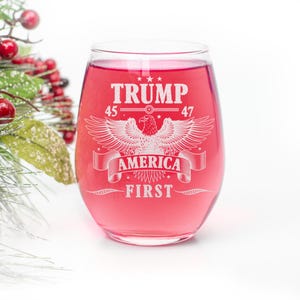 Puede incluir: Una copa de vino de vidrio transparente con un l&iacute;quido rojo en su interior. La copa tiene un dise&ntilde;o blanco con el texto "TRUMP 45 47 AMERICA FIRST" y un gr&aacute;fico de &aacute;guila.