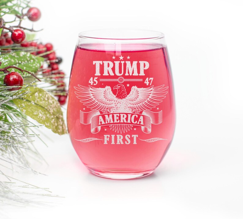 Puede incluir: Una copa de vino de vidrio transparente con un l&iacute;quido rojo en su interior. La copa tiene un dise&ntilde;o blanco con el texto "TRUMP 45 47 AMERICA FIRST" y un gr&aacute;fico de &aacute;guila.