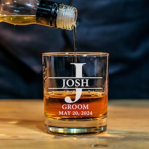 May include: A personalized whiskey glass with the name "Josh" and the text "Groom May 20, 2024" etched on it. Whiskey is being poured into the glass.
