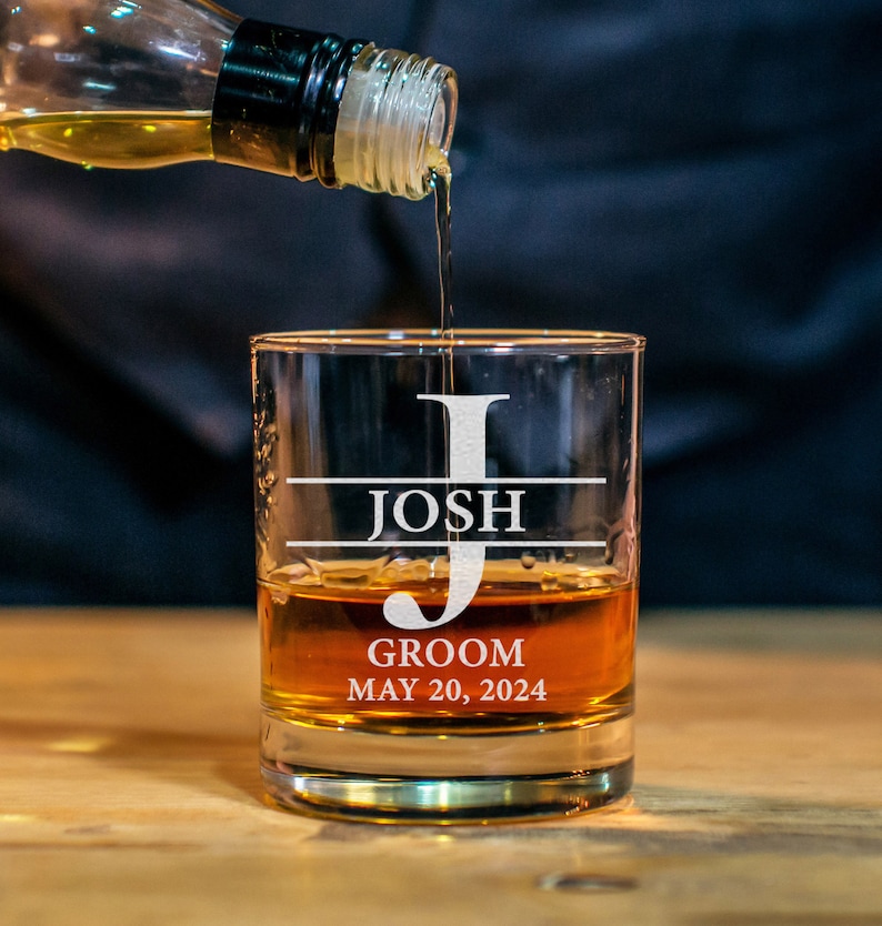 May include: A personalized whiskey glass with the name "Josh" and the text "Groom May 20, 2024" etched on it. Whiskey is being poured into the glass.