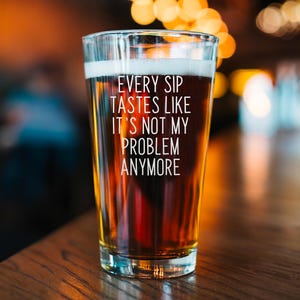 Pode incluir: Um copo de cerveja transparente cheio de cerveja &acirc;mbar com espuma branca. O copo tem o texto "EVERY SIP TASTES LIKE IT'S NOT MY PROBLEM ANYMORE" em branco. O copo est&aacute; sobre uma superf&iacute;cie de madeira.