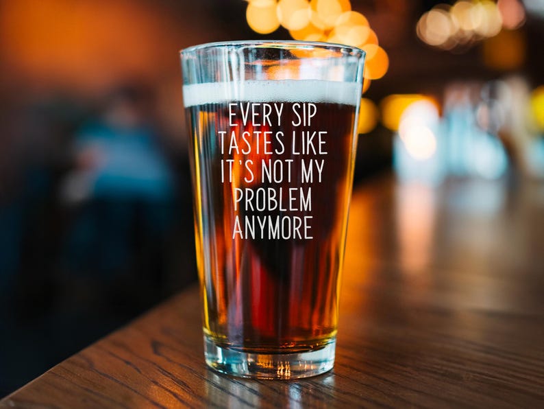 Pode incluir: Um copo de cerveja transparente cheio de cerveja &acirc;mbar com espuma branca. O copo tem o texto "EVERY SIP TASTES LIKE IT'S NOT MY PROBLEM ANYMORE" em branco. O copo est&aacute; sobre uma superf&iacute;cie de madeira.