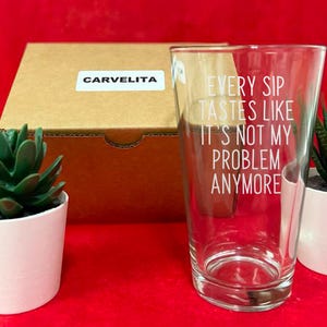 Pode incluir: Um copo transparente com o texto "EVERY SIP TASTES LIKE IT'S NOT MY PROBLEM ANYMORE" em letras brancas. O copo est&aacute; ao lado de uma caixa de cart&atilde;o com um adesivo "CARVELITA" e duas pequenas suculentas em vasos sobre uma superf&iacute;cie vermelha.