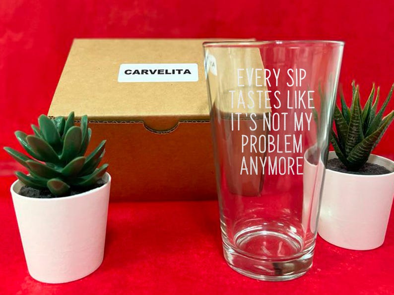 Pode incluir: Um copo transparente com o texto "EVERY SIP TASTES LIKE IT'S NOT MY PROBLEM ANYMORE" em letras brancas. O copo est&aacute; ao lado de uma caixa de cart&atilde;o com um adesivo "CARVELITA" e duas pequenas suculentas em vasos sobre uma superf&iacute;cie vermelha.