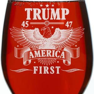 Puede incluir: Una copa de vino tinto con un dise&ntilde;o blanco que presenta un &aacute;guila con las alas extendidas, estrellas y el texto "TRUMP 45 47 AMERICA FIRST".