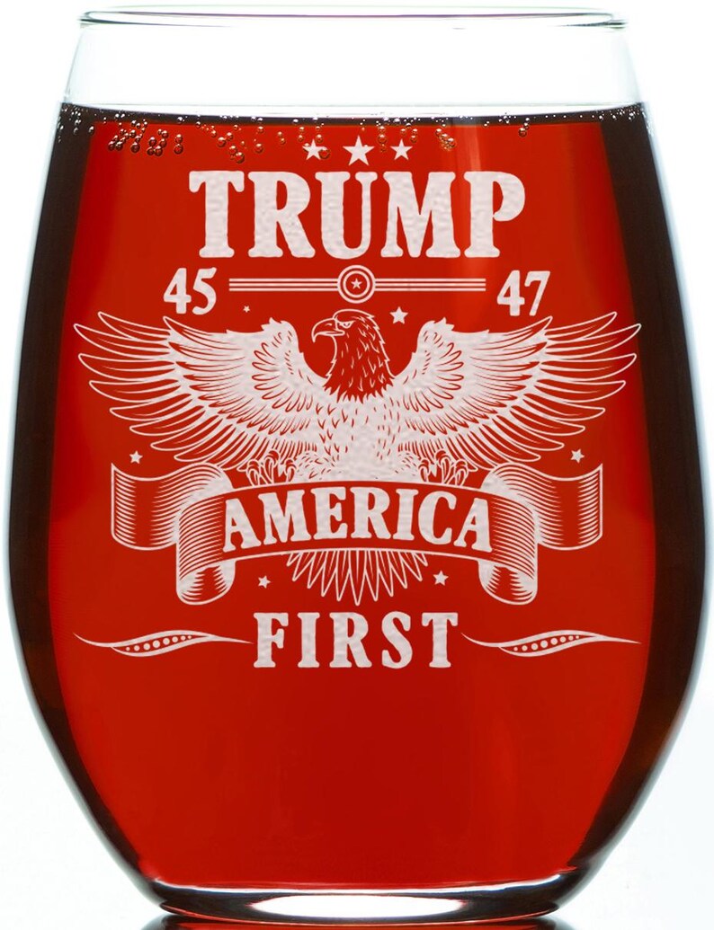 Puede incluir: Una copa de vino tinto con un dise&ntilde;o blanco que presenta un &aacute;guila con las alas extendidas, estrellas y el texto "TRUMP 45 47 AMERICA FIRST".
