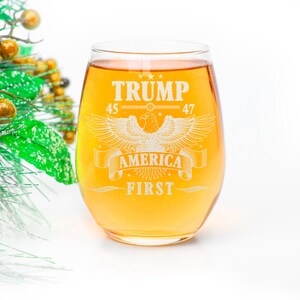 Puede incluir: Una copa de vino de vidrio transparente con un l&iacute;quido dorado en su interior. La copa tiene un dise&ntilde;o grabado de un &aacute;guila con las palabras "TRUMP 45 47 AMERICA FIRST" alrededor.