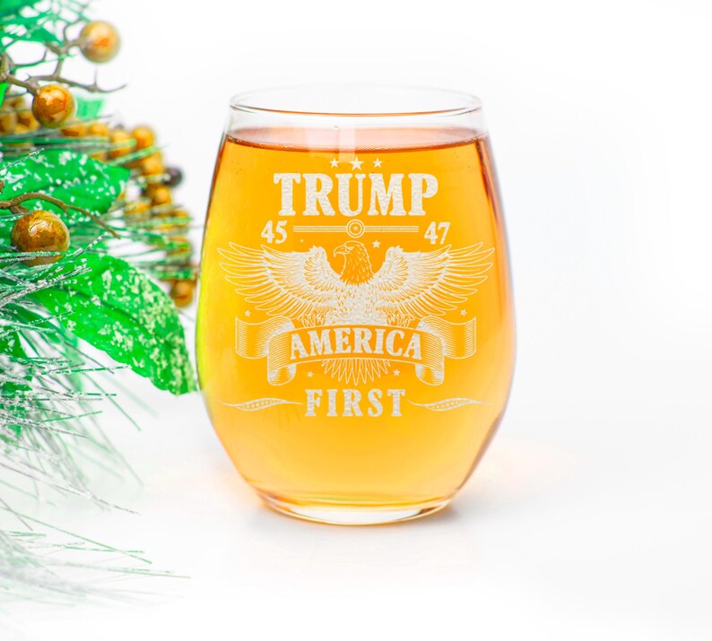 Puede incluir: Una copa de vino de vidrio transparente con un l&iacute;quido dorado en su interior. La copa tiene un dise&ntilde;o grabado de un &aacute;guila con las palabras "TRUMP 45 47 AMERICA FIRST" alrededor.