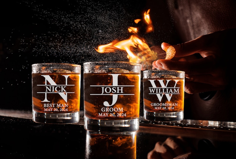 May include: Three personalized whiskey glasses with the names "Nick", "Josh", and "William" etched on them. The glasses are filled with amber liquid and have the words "Best Man", "Groom", and "Groomsman" printed below the names. The date "May 20, 2024" is printed below each title.