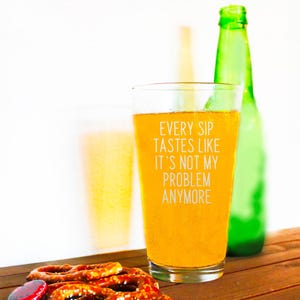 Pode incluir: Um copo de cerveja cheio de uma bebida dourada, com o texto "EVERY SIP TASTES LIKE IT'S NOT MY PROBLEM ANYMORE". Uma garrafa verde est&aacute; atr&aacute;s do copo. Pretzels em primeiro plano.
