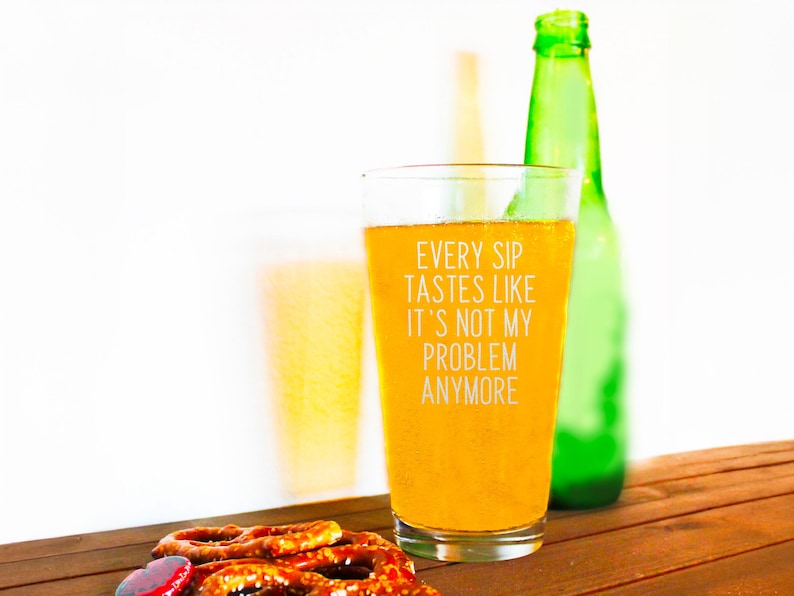 Pode incluir: Um copo de cerveja cheio de uma bebida dourada, com o texto "EVERY SIP TASTES LIKE IT'S NOT MY PROBLEM ANYMORE". Uma garrafa verde est&aacute; atr&aacute;s do copo. Pretzels em primeiro plano.
