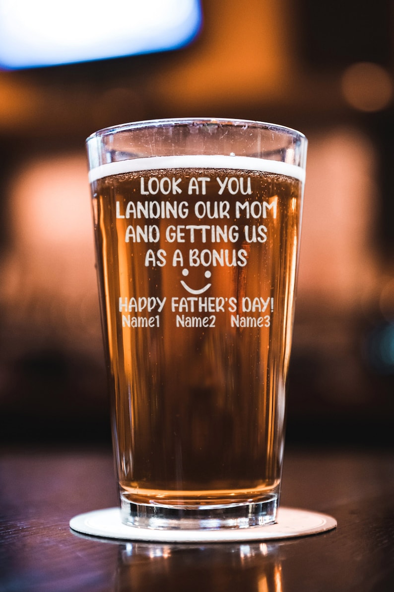 May include: A clear glass beer mug with a brown beer inside. The mug has an inscription that reads "Look at you landing our mom and getting us as a bonus. Happy Father's Day! Name1 Name2 Name3".