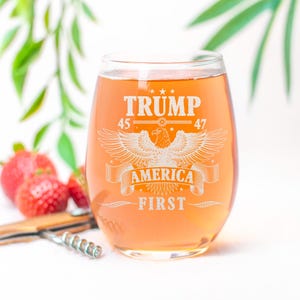 Puede incluir: Una copa de vino de vidrio transparente con un dise&ntilde;o blanco que dice "Trump 45 47 America First" con un gr&aacute;fico de &aacute;guila. La copa est&aacute; llena de un l&iacute;quido naranja claro.
