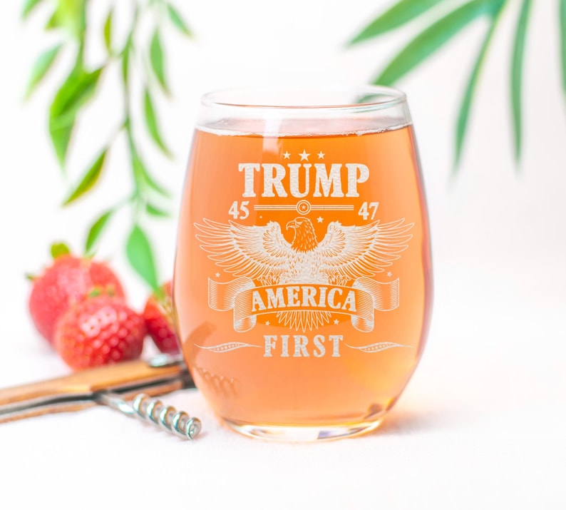 Puede incluir: Una copa de vino de vidrio transparente con un dise&ntilde;o blanco que dice "Trump 45 47 America First" con un gr&aacute;fico de &aacute;guila. La copa est&aacute; llena de un l&iacute;quido naranja claro.