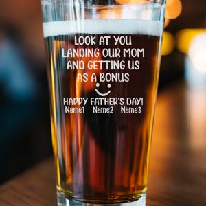 May include: A clear glass beer mug with a brown beer inside. The mug has a white inscription that reads "Look at you landing our mom and getting us as a bonus. Happy Father's Day! Name1 Name2 Name3".