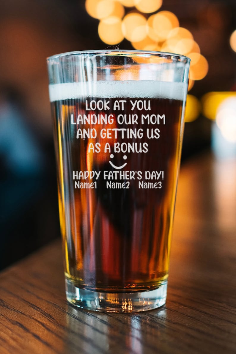 May include: A clear glass beer mug with a brown beer inside. The mug has a white inscription that reads "Look at you landing our mom and getting us as a bonus. Happy Father's Day! Name1 Name2 Name3".