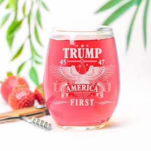 Puede incluir: Una copa de vino de vidrio transparente con un l&iacute;quido rojo en su interior. La copa tiene un dise&ntilde;o blanco con el texto "TRUMP 45 47 AMERICA FIRST" y un gr&aacute;fico de &aacute;guila.