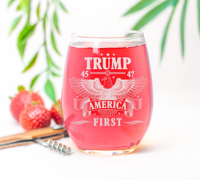 Puede incluir: Una copa de vino de vidrio transparente con un l&iacute;quido rojo en su interior. La copa tiene un dise&ntilde;o blanco con el texto "TRUMP 45 47 AMERICA FIRST" y un gr&aacute;fico de &aacute;guila.