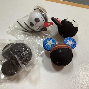 May include: A collection of four spherical ornaments featuring character designs. One is a white Jack Skellington head, one is a black mouse ear style, one is a black and blue patriotic style with glitter stars, and one is a plain black sphere in plastic.