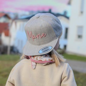 Könnte beinhalten: Hellgraue Baseballkappe mit dem Namen "Marie" in rosa Schrift gestickt. Die Kappe hat einen gebogenen Schirm und einen "Snapback"-Aufkleber. Die Person trägt einen beigen Pullover und einen floralen Schal.
