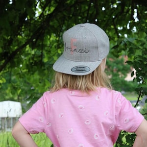 Könnte beinhalten: Eine hellgraue Snapback-Cap mit einem großen rosa Buchstaben F und dem in Schreibschrift gestickten Text Franzi auf der Vorderseite. Die Cap hat einen flachen Schirm mit einem runden Aufkleber darauf. Das Design ist mittig auf der Vorderseite platziert.