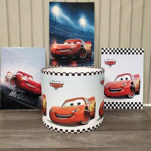 May include: A cylindrical lampshade featuring a repeating pattern of the red race car Lightning McQueen from Disney Pixar Cars, accented with a black and white checkered border. Three rectangular posters depicting the same character are displayed behind.