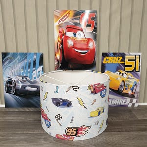 May include: A cylindrical lampshade featuring a repeating pattern of Lightning McQueen, Cruz Ramirez, and Jackson Storm race cars, checkered flags, lightning bolts, and the number 95. Three rectangular posters featuring the same characters are behind it.