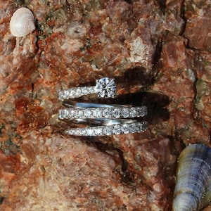 May include: Three silver rings with a large diamond in the center, set on a rough, brown, rocky surface.