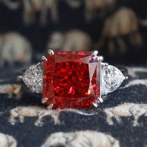 Può includere: Un anello d'argento con una grande pietra rossa di forma quadrata circondata da pietre più piccole e trasparenti.