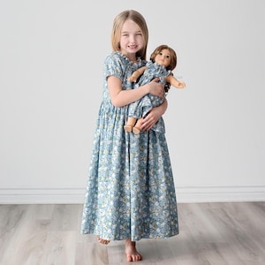 May include: A young girl in a blue floral dress holds a doll wearing a matching dress. The doll has long brown hair and blue eyes.