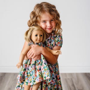 Può includere: Una bambina con un vestito floreale tiene in braccio una bambola che indossa un vestito floreale abbinato. La bambina e la bambola sorridono e guardano la fotocamera.