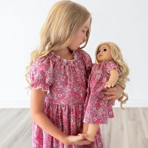 May include: A young girl in a pink floral dress holds a matching doll. The dresses feature a pink background with a white and gray floral pattern. Both dresses have short sleeves with white trim and a gathered neckline.