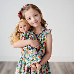 May include: A young girl in a floral print dress is holding a doll wearing a matching dress. The girl has long blonde hair and is smiling. The doll has blonde hair and blue eyes.