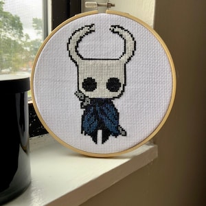 May include: A cross-stitch embroidery of a character from the video game Hollow Knight. The character is white with large black eyes, a dark blue cloak, and a sword. The embroidery is set in a round wooden hoop.