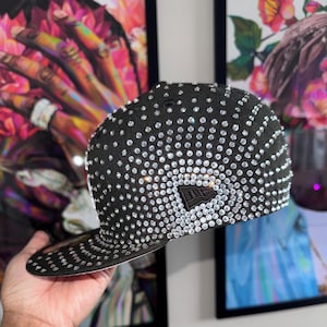 Pode incluir: Um bon&eacute; de beisebol preto com um grande padr&atilde;o de diamantes de imita&ccedil;&atilde;o brancos em forma de diamante. O bon&eacute; tem uma viseira preta e um logotipo New Era preto na frente.